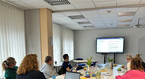 Teilnehmende des Mentorentreffens sitzen in großem Raum am Tisch und schauen nach vorne auf die PowerPoint-Präsentation.