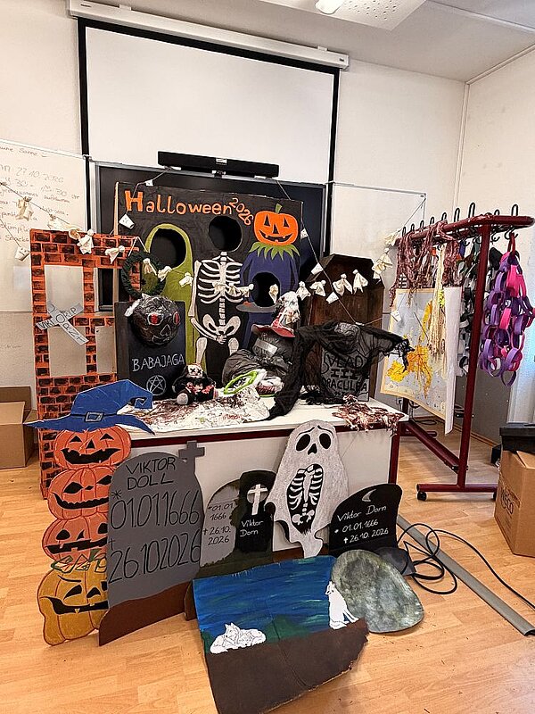Halloween-Deko im Klassenzimmer: Auf einem Tisch sind ein Skelett, ein Geisterkostüm, eine Zombie-Figur, selbstgebastelte Grabsteine u.a. mit der Aufschrift ‚Viktor Doll' und Grusel-Kürbisse aus Pappe arrangiert. Im Hintergrund hängt ein Banner mit ‚Halloween 2026', dazu Spinnennetz und Girlanden an den Wänden.