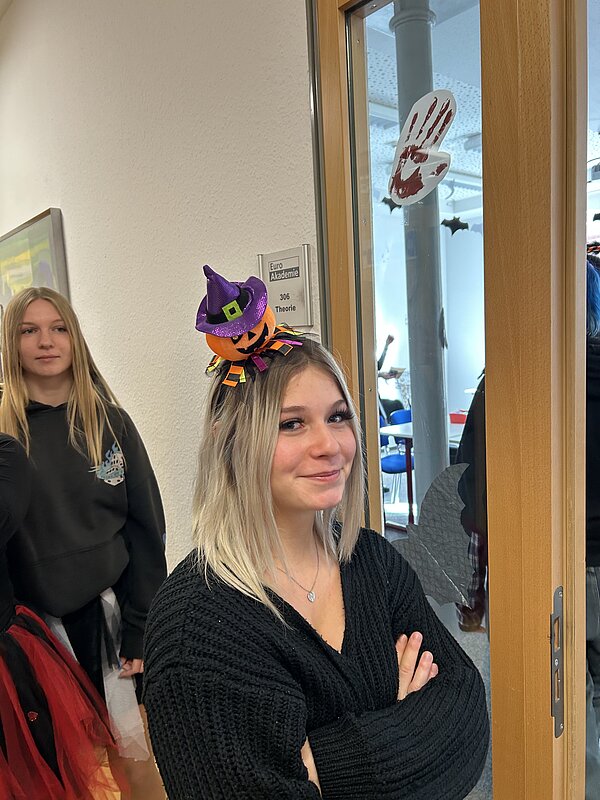 Teilnehmerin mit Mini-Hexenhut-Haarreif lächelt vor einer Tür mit der Beschriftung „Euro Akademie – Theorie“; Blut-Handabdruck als Deko am Glas.