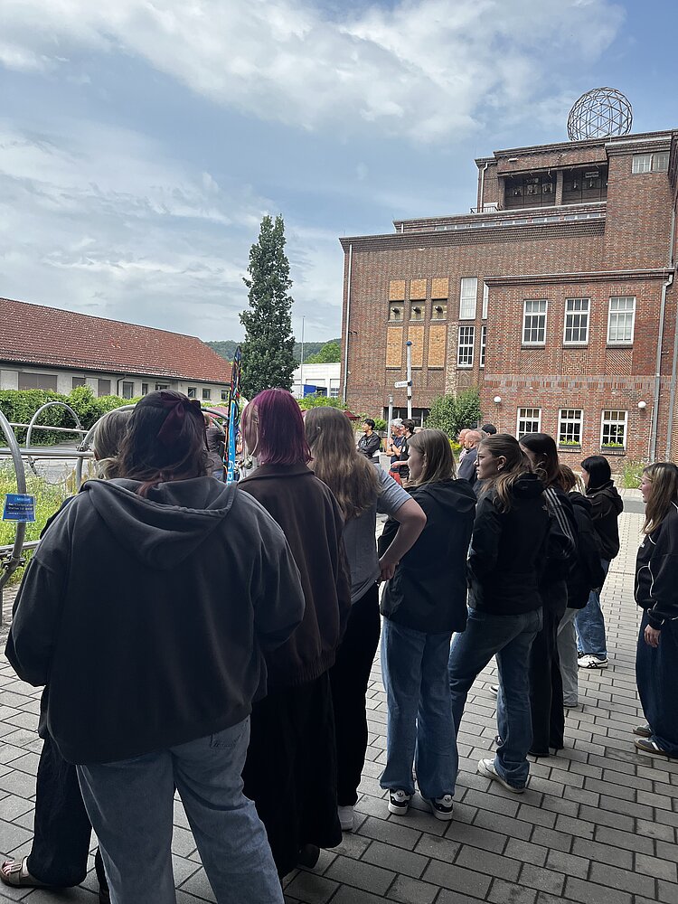 Auf einem Platz vor einem Gebäude mit braunroten Ziegeln steht eine Gruppe Schülerinnen und Schüler und schaut nach links.