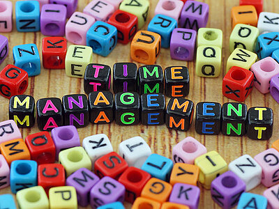 Das Bild zeigt bunte Buchstabenwürfel auf einer Holzoberfläche. In der Mitte sind die Worte TIME MANAGEMENT aus den Würfeln geformt. 
