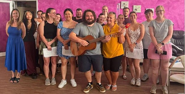 Vor einer pinken Wand steht eine Gruppe lächelnder Menschen. Ein Mann in der Mitte hält eine Gitarre. Eine Frau hält eine Rassel. Mehrere Frauen halten Liedtexte in den Händen.