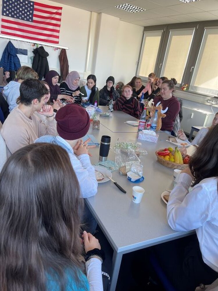Eine größere Gruppe von Menschen sitzt um einen rechteckigen Tisch in einem hellen Raum. Der Tisch ist mit Frühstücksartikeln wie Obst, Brot und Getränken gedeckt. Im Hintergrund hängt eine amerikanische Flagge an der Wand, und an der Garderobe sind Jacken aufgehängt. Die Anwesenden unterhalten sich, essen oder schauen interessiert in die Runde. Große Fenster lassen viel Tageslicht in den Raum. Die Szene vermittelt eine gemütliche und gemeinschaftliche Stimmung.