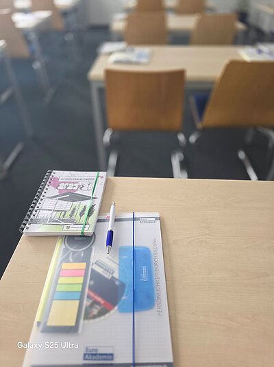 Auf einem Tisch in einem Klassenzimmer liegen Unterrichtsmaterialien, wie einem Schülerkalender, einem durchsichtigen Hefter, der einen Stift, bunte Post-its und einen Schreibblock mit dem Euro Akademie Logo enthält.