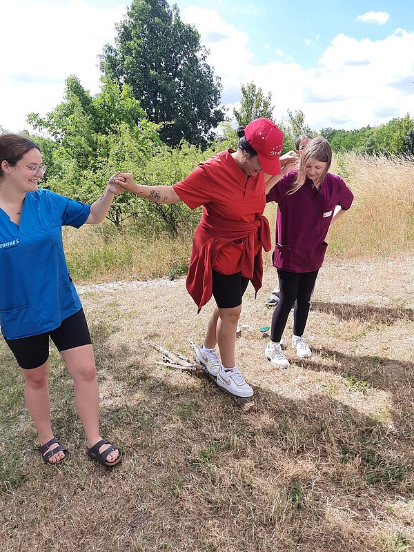 Ein Sinneskreis am Goitzsche-Strand: Lernen, Fühlen, Erleben! Drei Frauen bei einer Balance-Übung im Freien. Eine Frau balanciert über einen Ast, die anderen beiden reichen ihr die Hand zur Unterstützung.
