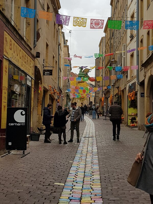 Schulfahrt nach Metz Blick in eine schöne kleine Gasse in der Altstadt von Metz, mit Kopfsteinpflaster und bunten Girlanden, die oben zwischen den Häusern angebracht sind und Menschen flanieren.