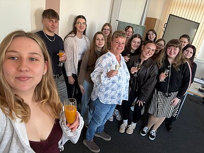 Gruppen-Selfie in einem Klassenraum: Eine Gruppe junger Erwachsener und eine Lehrkraft stehen zusammen, lächeln in die Kamera und halten Gläser mit Saft bzw. Sekt zum Anstoßen.