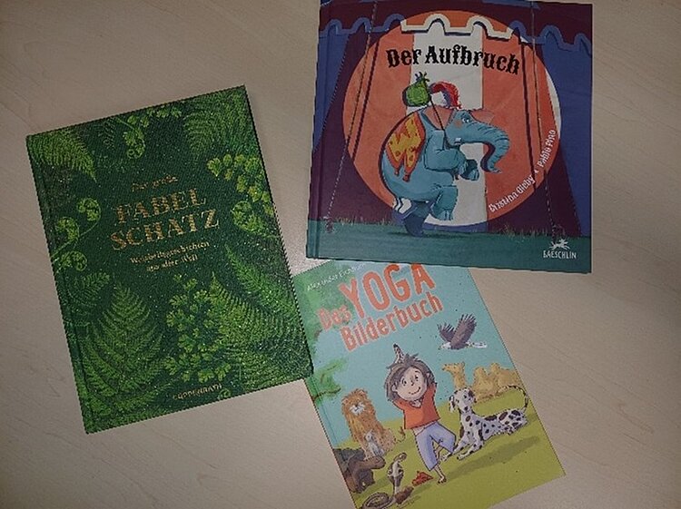 Drei farbenfrohe Kinderbücher liegen auf einem hellen Holztisch. Zu sehen sind: das grüne Buch Fabelschatz – Wundersame Fabeln aus aller Welt, das blau-rote Buch Der Aufbruch mit einer Illustration eines Elefanten, sowie das hellblaue Yoga-Bilderbuch mit einem fröhlichen Kind in Yogaposition und verschiedenen Tieren. Die Bücher wurden von Auszubildenden der Euro Akademie Dessau zum Bundesweiten Vorlesetag genutzt, um Kinder für Sprache, Bewegung und Geschichten zu begeistern.