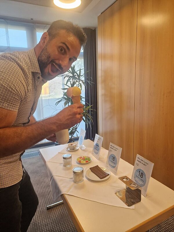 Ein Mann in einem hellen, gemusterten Hemd hält ein Eis in einer Waffel in der Hand und schaut dabei euphorisch in die Kamera. Auf dem Tisch vor ihm stehen verschiedene Süßigkeiten wie Schokolade und Gummibärchen sowie eine Tasse mit Cappuccino. Im Hintergrund sind ein Fenster, eine Zimmerpflanze und eine Holzvertäfelung zu sehen.