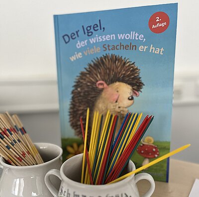 Kinderbuch mit dem Titel „Der Igel, der wissen wollte, wie viele Stacheln er hat“ steht aufrecht auf einem Tisch. Davor zwei Becher mit bunten Holzstäbchen, ähnlich wie bei einem Mikado-Spiel.