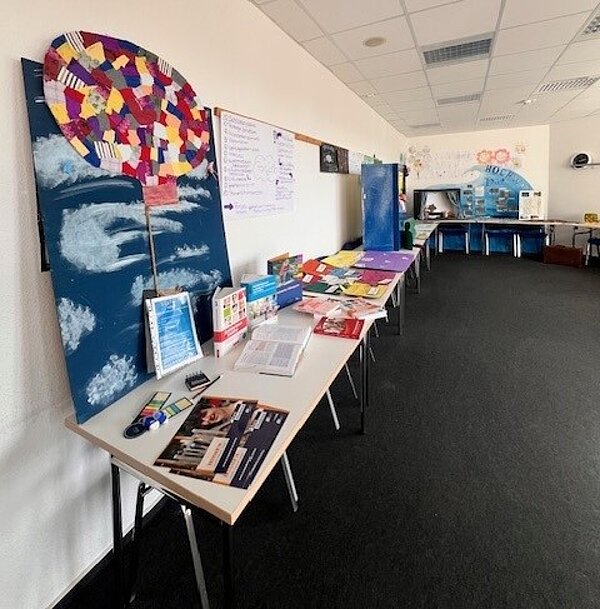 Ein Klassenzimmer oder Gemeinschaftsraum mit mehreren Tischen, die entlang der Wand aufgestellt sind. Auf den Tischen liegen Informationsmaterialien, Broschüren, Hefte, Stifte und andere Bastelutensilien. An der Wand hängt ein großes, buntes Kunstwerk, das einen Heißluftballon in einem Himmel mit Wolken darstellt. Im Hintergrund sind weitere Materialien und ein Whiteboard mit Notizen zu sehen.
