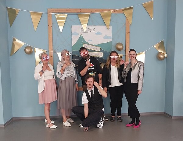 Herzlichen Glückwunsch! Sechs Absolventinnen und Absolventen posieren bei Abschlussfeier für ein Gruppenfoto. Sie tragen festliche Kleidung, stehen vor einer pastellblauen Wand, an der Girlanden mit Wimpeln aus Gold aufgehängt sind und halten sich Foto-Booth-Requisiten vor das Gesicht.