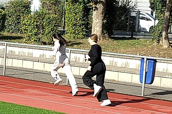 Zwei Personen laufen auf einer roten Tartanbahn in einem Sportstadion. Im Hintergrund sind Bäume und ein Zaun zu sehen.