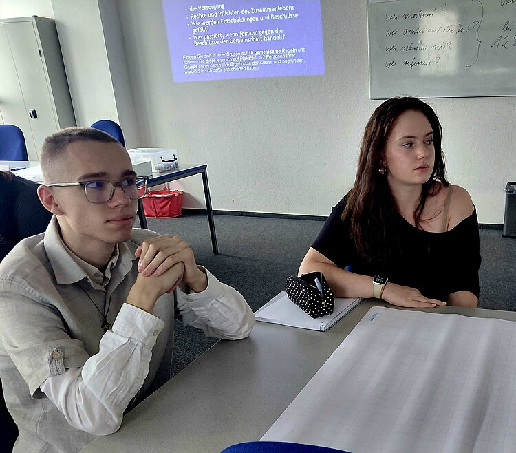 In einem Klassenzimmer sitzen ein Schüler und eine Schülerin an einem Tisch. Sie schauen nach rechts auf etwas, was außerhalb des Bildausschnittes liegt. Auf dem Tisch vor ihnen liegen Papier und ein Mäppchen. Im Hintergrund sind eine Tafel und eine Power-Point-Präsentation an der Wand zu sehen.