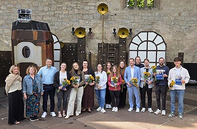 Eine Gruppe von Menschen steht vor einer künstlerisch gestalteten Bühnenkulisse aus Holz und Metall vor einer Steinmauer. Viele von ihnen halten Zertifikate sowie Sonnenblumensträuße.