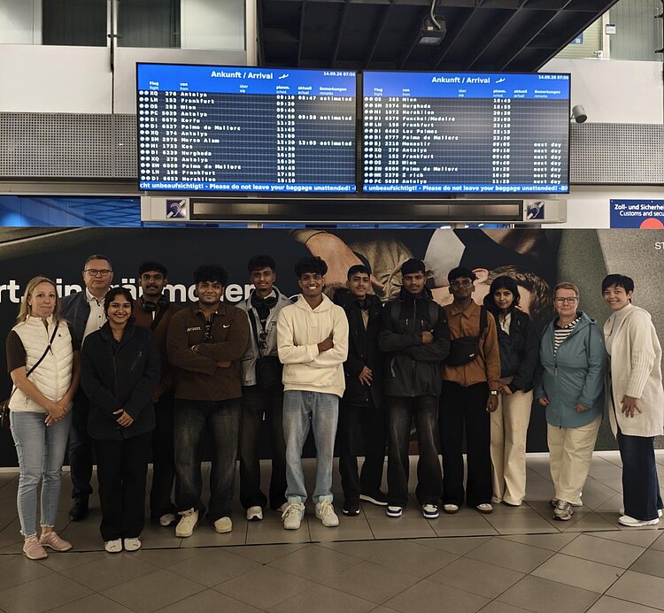Eine größere Gruppe von 13 Personen steht in einer Reihe in einem Flughafenterminal. Im Hintergrund sind zwei digitale Anzeigetafeln mit Fluginformationen (Ankunft/Arrival) zu sehen. Die Gruppe besteht aus einer vielfältigen Mischung von Personen, die lächeln oder entspannt posieren. Einige tragen Jacken, Rucksäcke oder andere Reiseaccessoires.