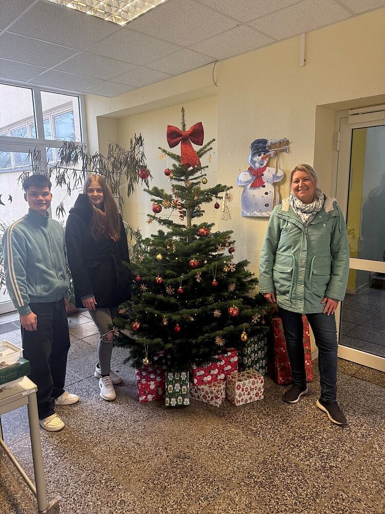 Drei lächelnde Personen – eine junge Frau, ein junger Mann und eine ältere Frau – stehen nebeneinander in einem Innenraum. Sie posieren vor einem geschmückten Weihnachtsbaum, der mit roten Kugeln, goldenen Sternen, einer großen roten Schleife an der Spitze und Lichtern dekoriert ist. Unter dem Baum liegen mehrere bunt verpackte Geschenke. Im Hintergrund hängt eine Wanddekoration mit einem Schneemann. Die ältere Frau trägt einen hellen, grünlichen Mantel und ein graues Schal-Tuch, die junge Frau einen dunklen Mantel und der junge Mann ein helles Sweatshirt mit Streifen an den Ärmeln. Der Raum wirkt freundlich und festlich.