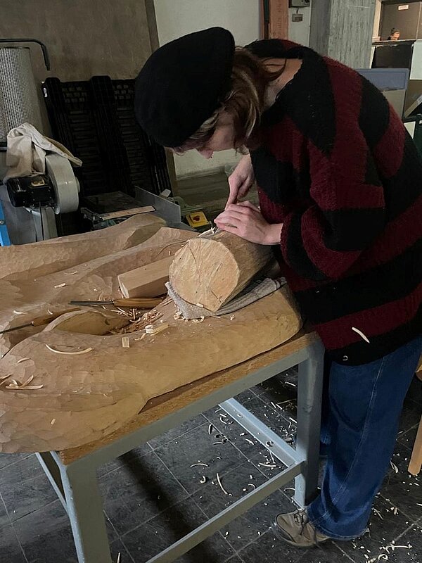 Eine Person mit schwarzer Mütze arbeitet an einem Arbeitstisch an einem Holzstück, welches auf großen bearbeiteten Holzstücken liegt. Ein graues Tuch schützt die Auflage. Auf Tisch und Boden liegen Holzspäne.