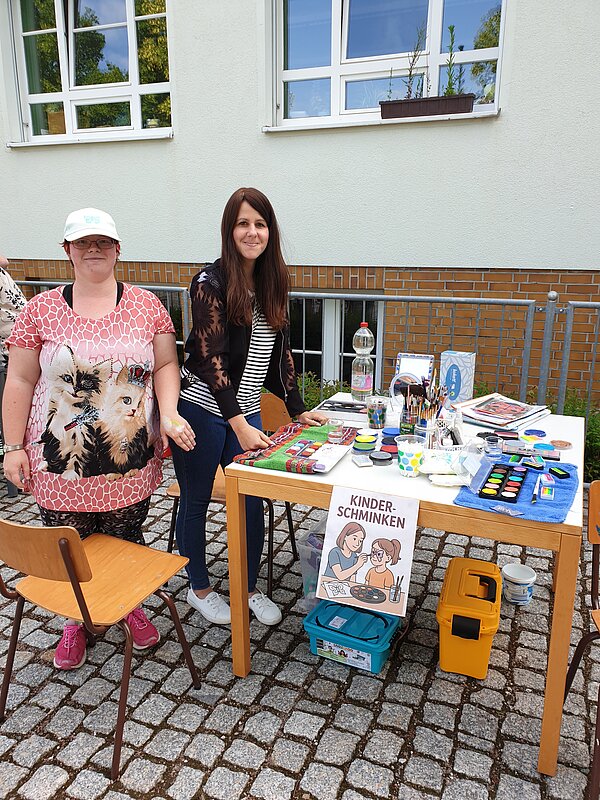 Ein erlebnisreicher Vormittag voller Spiel, Kreativität und Gemeinschaft Zwei Frauen an einem Tisch mit Schminkutensilien, auf dem ein Schild „Kinderschminken“ steht, für ein kreatives Mitmachangebot.