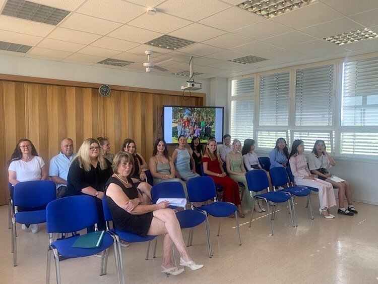 Herzlichen Glückwunsch! Ein Gruppenfoto von mehreren Personen, darunter junge Frauen und einige ältere Erwachsene, die in einem Seminarraum mit blauen Stühlen sitzen. Im Hintergrund ist ein Bildschirm zu sehen, der ein Bild einer Gruppe im Freien zeigt. Die Personen sind festlich angezogen. Sie lächeln oder schauen entspannt, einige halten Unterlagen. Der Raum hat große Fenster mit Jalousien und eine Holzwand mit einer Uhr.