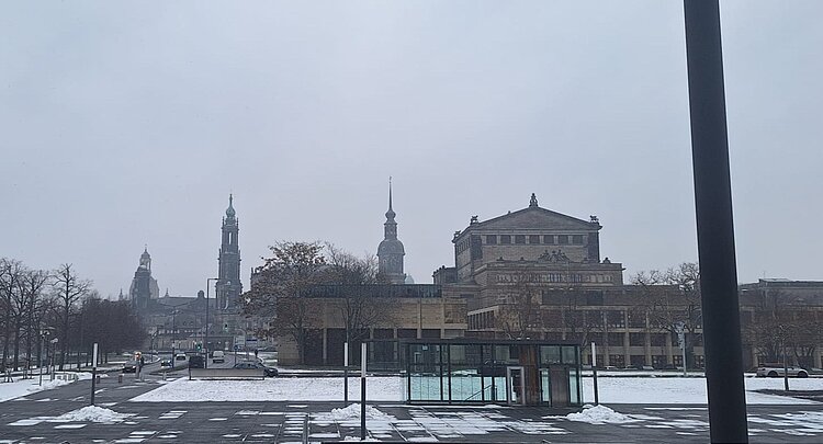 Blick auf einen verschneiten, weitläufigen Platz in Dresden mit Straßen und wenigen Autos im Vordergrund, dahinter mehrere historische Gebäude mit Türmen unter einem grauen Winterhimmel.