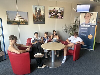 In der Lounge der Euro Akademie Magdeburg sitzen fünf Jugendliche auf roten Sofas und Sesseln und sind vertieft in ihre Lektüre. Im Hintergrund steht ein blaues Roll-up der Schule, auf der die angebotenen Ausbildungen festgehalten sind, darüber thront ein digitaler Bildschirm über dem Geschehen, an der Wand hängen großformatige Bilder von Sehenswürdigkeiten der Stadt.