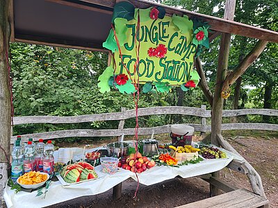 Ein überdachter Holztisch im Freien, dekoriert mit einem großen Schild „Jungle Camp Food Station“. Auf dem Tisch stehen Wassermelonenstücke, Chips, frisches Obst, Gummibärchen, Wasserflaschen und weitere Snacks.