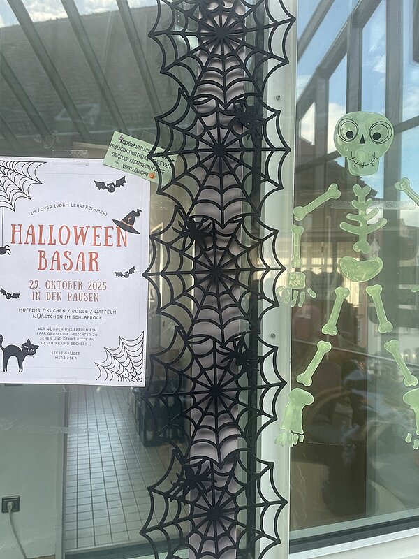 Halloween-Kuchenbasar in der Euro Akademie und den Euro-Schulen in Meißen Poster des Halloweenbasars hängt am Eingangsbereich am Fenster, dekoriert mit Spinnennetzen.