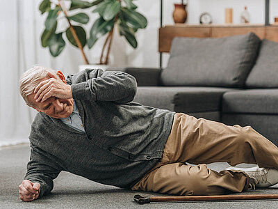 Auf dem Bild liegt ein älterer Mann am Boden neben einem Gehstock. Er trägt einen grauen Pullover, hält sich den Kopf und wirkt unwohl. Im Hintergrund sind ein graues Sofa, ein Regal mit verschiedenen Gegenständen und eine Topfpflanze zu sehen.