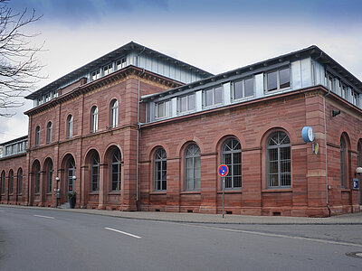 Ein zweistöckiges Gebäude aus rotem Sandstein mit einer Mischung aus historischen und modernen architektonischen Elementen. Das Gebäude hat große, rundbogige Fenster im Erdgeschoss und rechteckige Fenster im Obergeschoss. Die Fassade ist mit dekorativen Elementen verziert, und das Dach ist flach mit einer modernen Metallkonstruktion. Das Gebäude steht an einer ruhigen Straße mit einem Gehweg und einem Baum im Vordergrund.