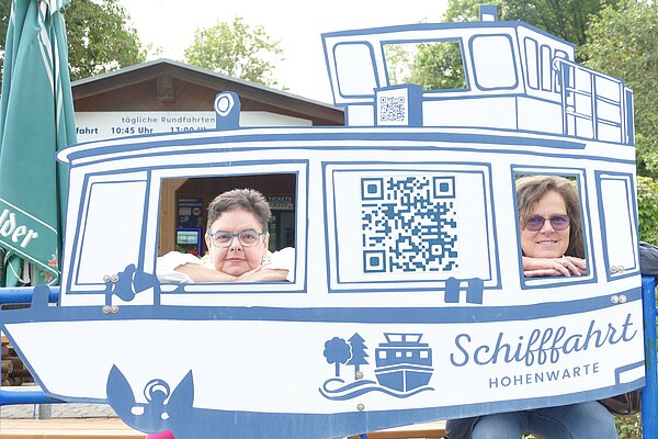 Zwei Frauen schauen durch die Fenster einer großen Fotowand in Form eines Schiffes mit der Aufschrift „Schifffahrt Hohenwarte“.