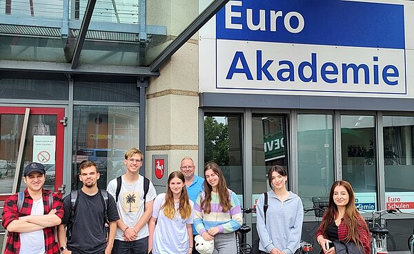 Schülerinnen und Schüler in sommerlicher Kleidung mit Rucksäcken stehen vor einem Gebäude mit dem blau-weißen Schriftzug Euro Akademie.