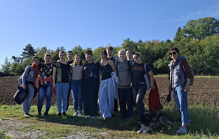 Zwölf Personen posieren lächelnd auf einem Grasweg, eine Person hält einen angeleinten Hund; dahinter ein gepflügtes Feld mit Bäumen unter blauem Himmel.
