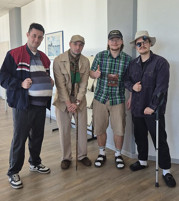 Vier Männer posieren verkleidet als Rentner mit typischen Accessoires wie Gehstock, Bauchtasche, Sonnenhut und gemütlicher Kleidung. Sie stehen lachend im Flur der Schule.