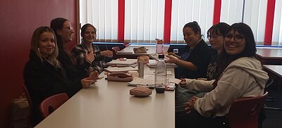 Mehrere Schülerinnen sitzen nach dem Unterricht gemeinsam an einem Tisch im Klassenraum, lachen in die Kamera und haben Hefte, Stifte und Getränke vor sich liegen.