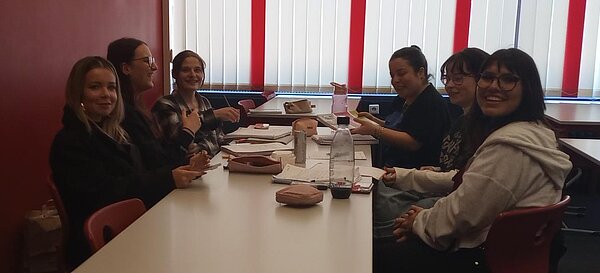 Mehrere Schülerinnen sitzen nach dem Unterricht gemeinsam an einem Tisch im Klassenraum, lachen in die Kamera und haben Hefte, Stifte und Getränke vor sich liegen.