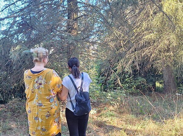 Zwei Frauen stehen in einem bewachsenen Waldstück und betrachten gemeinsam die Pflanzen und Bäume.