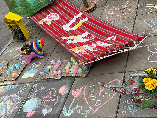 Auf Steinplatten sind zwischen einer Hängematte, einer bunten Schweinefigur und Blumen mit Kreide verschiedene Bilder und Texte aufgemalt.