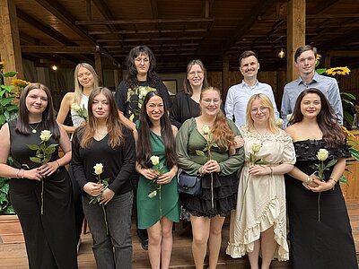 In festlicher Kleidung haben sich die Absolvent*innen für ein Gruppenfoto aufgestellt. In den Händen halten sie alle eine langstielige weiße Rose.