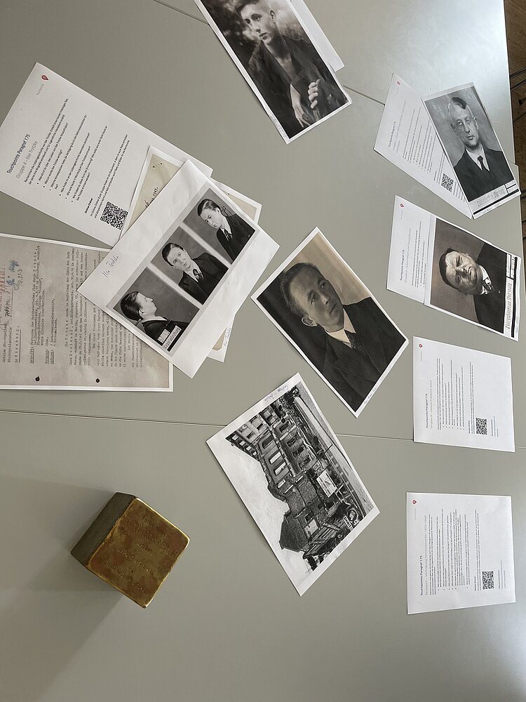 Historische Fotos, Biografien und Dokumente sind auf einem Tisch für die Spurenwerkstatt ausgebreitet. Neben den Materialien liegt ein goldfarbener Erinnerungsstein, der an ein Opfer der nationalsozialistischen Verfolgung erinnert.