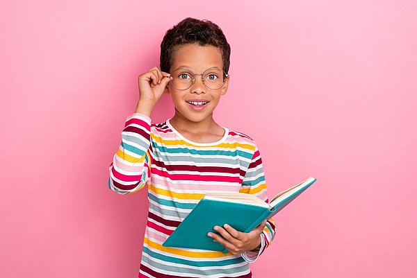 Kinderbuchklassiker im Unterricht kritisch beleuchtet · Teil 4 Ein kleiner Junge mit dunklen, kurzen Haaren und einem geringelten Langarmshirt steht vor einer rosafarbenen Wand. Er hält in der linken Hand ein Buch, mit der rechten Hand greift er seine Nickelbrille am Gestell.