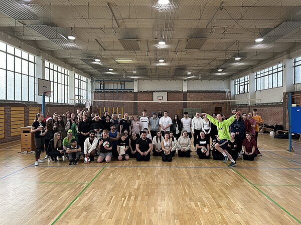 Gruppe aus Teilnehmenden des Volleyballturniers posiert in der Sporthalle, einige halten Volleybälle, vorne springt eine Person im neongrünen Shirt begeistert in die Luft.