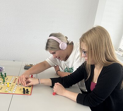 Zwei Schülerinnen sitzen an einem Tisch und spielen "Mensch ärgere dich nicht". Eine der beiden trägt einen Kopfhörer, während sie gleichzeitig ihre Figuren zu bewegen versuchen.