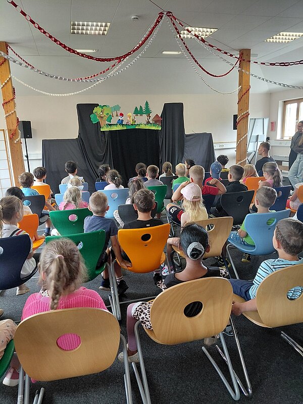 Kinder sitzen auf bunten Stühlen in einem Raum und schauen auf eine kleine Bühne, auf der ein Puppentheater mit Figuren und Kulisse aufgebaut ist. Über ihnen hängen rote und weiße Papiergirlanden. Die Kinder sind von hinten zu sehen und blicken aufmerksam nach vorne.