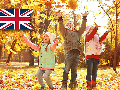 Drei fröhliche Kinder spielen im Herbstlaub, umgeben von gelben und orangefarbenen Blättern. Die britische Flagge ist oben links zu sehen.