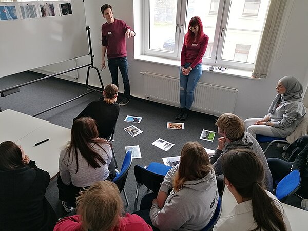 Die beiden Workshopleiter*innen stehen am Flipchart und referieren zumThema Migration und Flucht, während die Auszubildenden auf blauen gepolsterten Stühlen sitzen und zuhören. 
