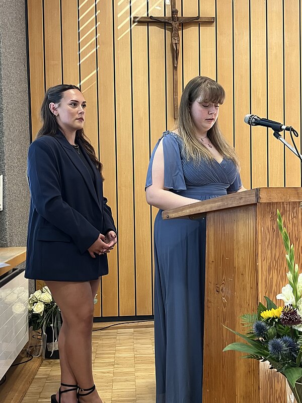 An einem Rednerpult aus Holz stehen zwei junge Frauen in Kleidern. Im Vordergrund steht ein Blumenstrauß. Hinten an der Holzwand hängt ein Kreuz mit Jesusfigur.