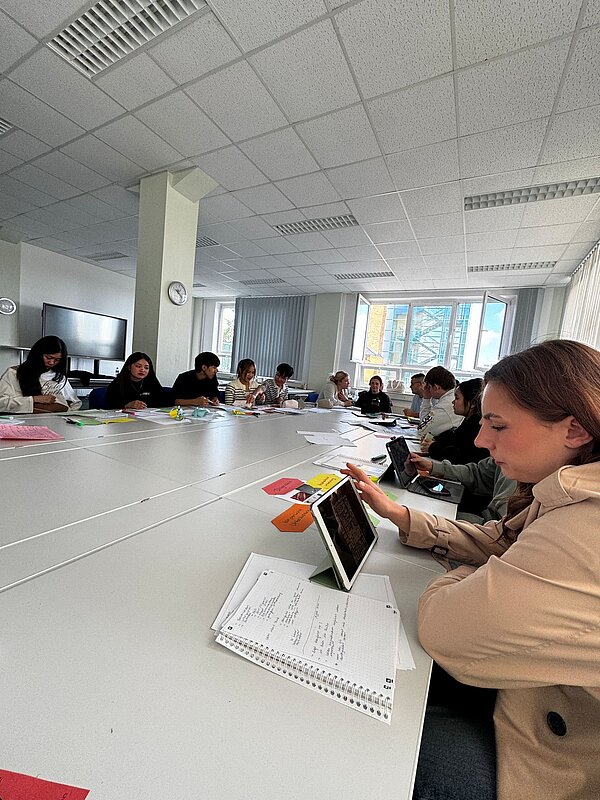 Eine größere Lerngruppe sitzt in einem hellen Klassenraum an einem langen Tisch. Die Schülerinnen und Schüler arbeiten mit Notizen, Tablets und farbigen Lernmaterialien zusammen.