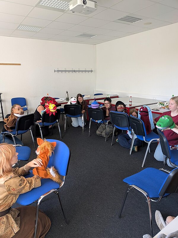 Die Azubis sitzen in einem hellen Raum auf blauen Stühlen im Kreis und spielen mit ihren verschiedenen bunten Handpuppen aus Plüsch im Rahmen des Workshops.