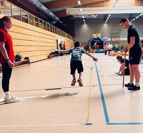 Kind springt in einer Turnhalle zwischen zwei betreuenden Personen über eine markierte Linie, im Hintergrund sind weitere Kinder, Eltern und eine Hüpfburg zu sehen.